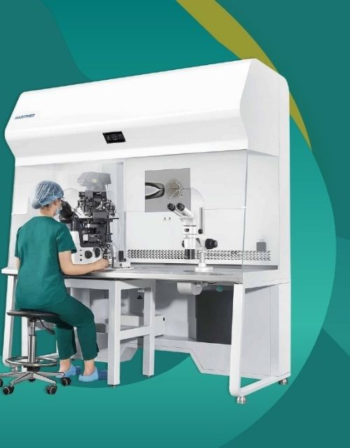 Embrióloga con uniforme quirúrgico trabajando dentro de una estación de trabajo para FIV (Fecundación In Vitro) marca Haromed. La profesional está sentada y utiliza un microscopio integrado en el ambiente estéril de la cabina de flujo laminar.