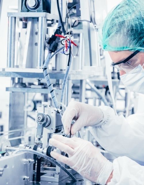 SERVICIO TECNICO. "Técnico especializado en una sala limpia, equipado con traje de protección estéril, gafas y guantes, realizando un ajuste de precisión en una máquina de una línea de producción farmacéutica o de dispositivos médicos.